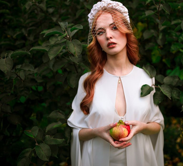 Bride in white wedding dress and bridal cape wearing white headband with net stands holding apple