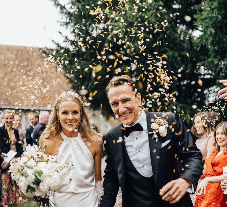 Bride in halterneck Halfpenny London wedding dress and bespoke floral headband holding white and green bridal bouquet walks through churchyard holding hands with groom in black tie as guests throw confetti