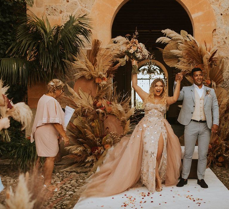 Bride raises her hands in celebration as she walks with groom on her wedding day