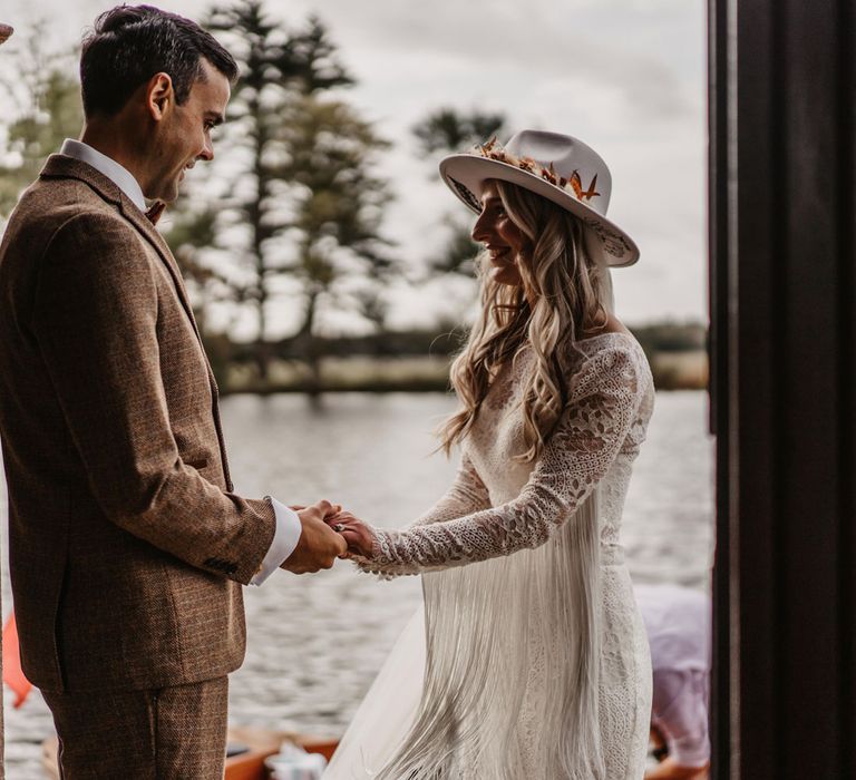 Groom in brown tweed suit holds hands with bride in two piece wedding dress with tassels and hat by lake before wedding