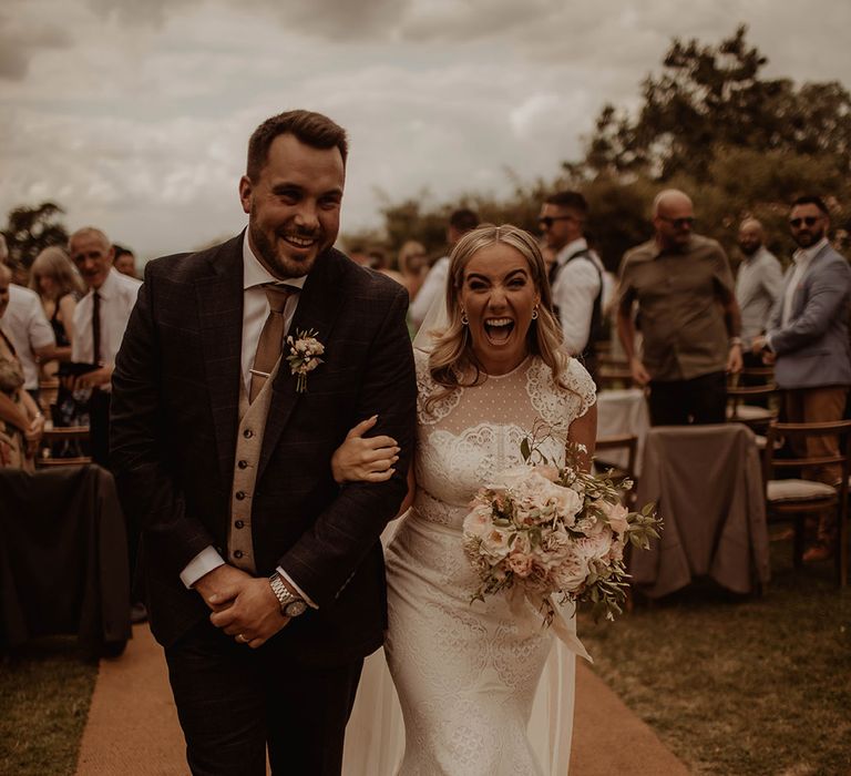 Bride in a lace cap sleeve wedding dress and groom in a three-piece suit descending up the aisle as husband and wife