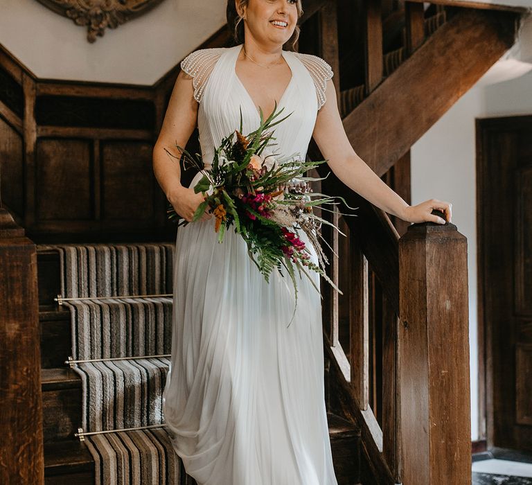 Bride wears wedding gown with capped sleeves and embellishment whilst holding Autumnal floral bouquet