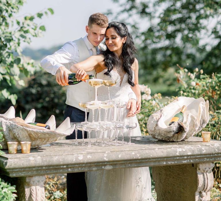 Bride & groom pour champagne into glass tower on their wedding day outdoors as the sun shines around them