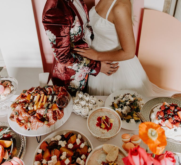 Two brides in a vervet jacket and short wedding dress embracing at the food table provided by Baba Ganoush