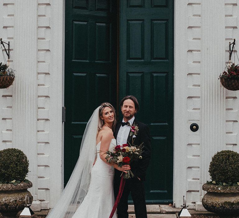 Bride & groom stand in front of green door on their wedding day as bride wears Helen Martin Veil and Suzanne Neville wedding gown