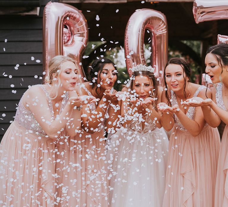 Bride and bridesmaids blow some confetti at the camera in front of rose gold balloons