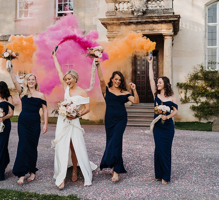 Bride in front slit wedding dress and bridesmaids in navy blue dresses hold up orange and pink smoke bombs