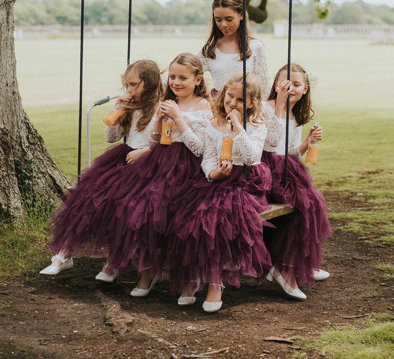 Flower girls in purple and lace dresses sit together on swing attached to a tree drinking J2O's 