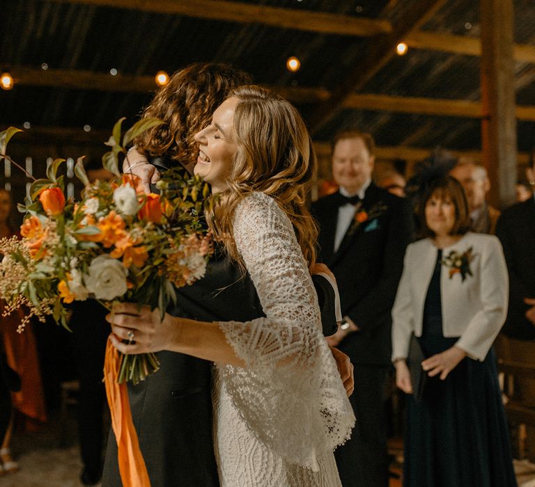 Groom in black tie hugs bride in lace Rosa Clara wedding dress with bell sleeves holding orange autumnal bouquet with ribbon 