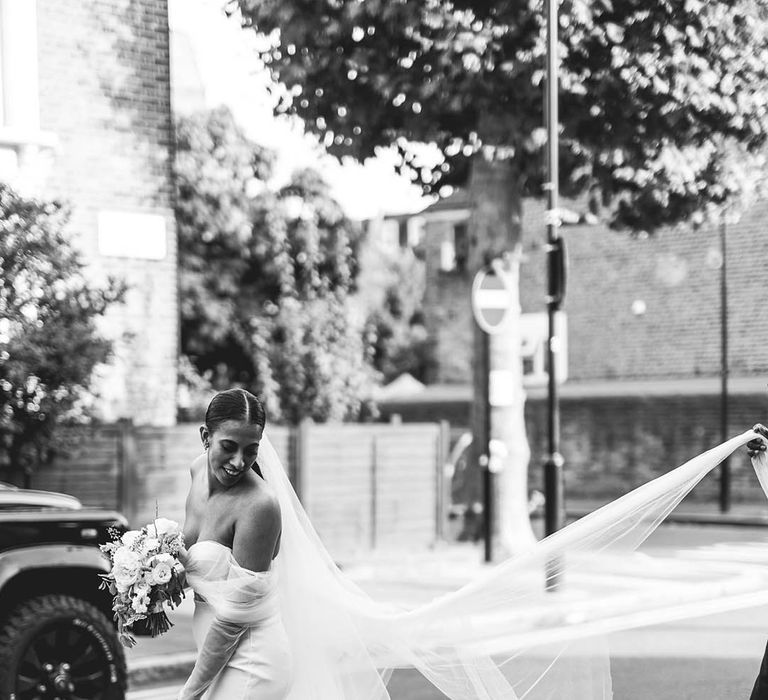 Groom helps lift up the bride's veil as she walks along the street in square toe heels and off the shoulder dress