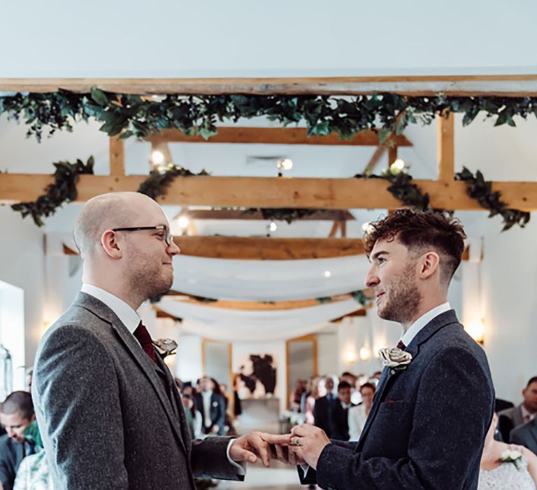 Groom in dark blue three piece wedding suit puts ring on groom in grey suit's finger during their ceremony at Southend Barns