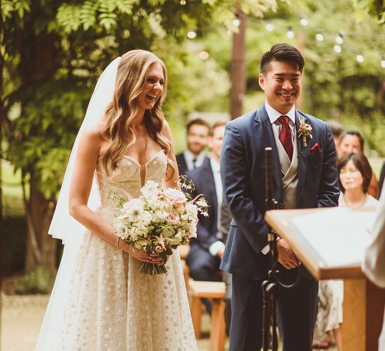 Smiling bride in Pronovias wedding dress stands at the altar with the groom in a blue suit for their outdoor wedding ceremony