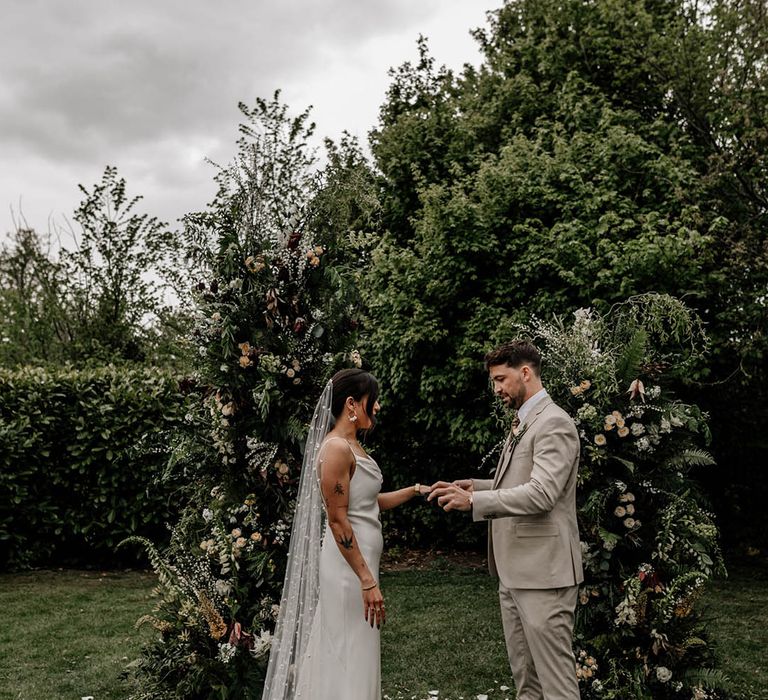 Groom in a beige wedding suit exchanging vows with his bride in a slip wedding dress and pearl veil at their outdoor wedidng ceremony with column wedding flowers 