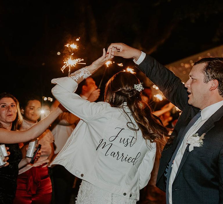 Groom spins around bride wearing white denim jacket reading 'Just Married' as they have their sparkler send-off 