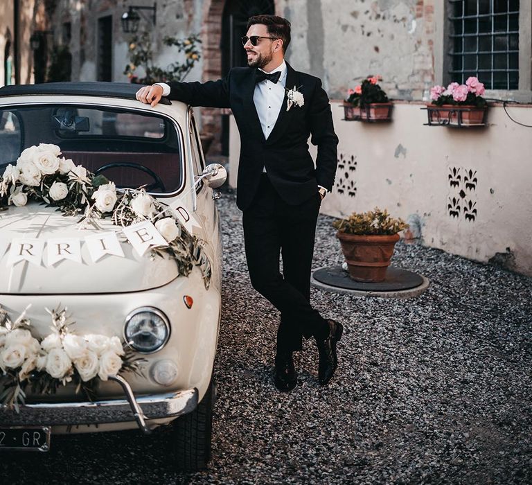 Groom in a tuxedo and sunglasses standing next to a fiat 500 decorated car at his black and white destination wedding 