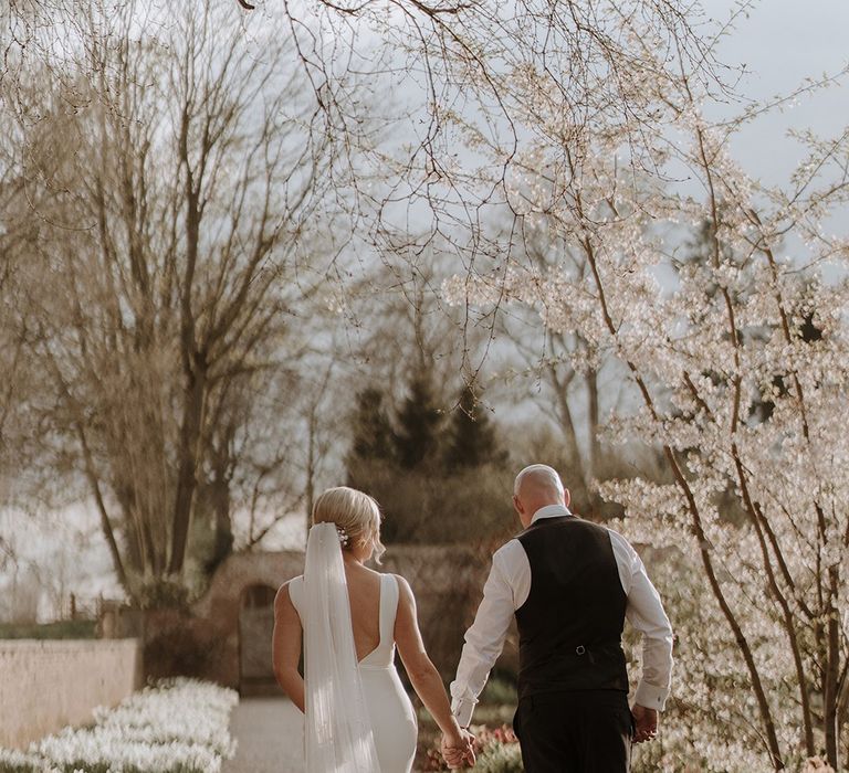 Bride in button back wedding dress walks holding hands with groom in black tie for their spring wedding 