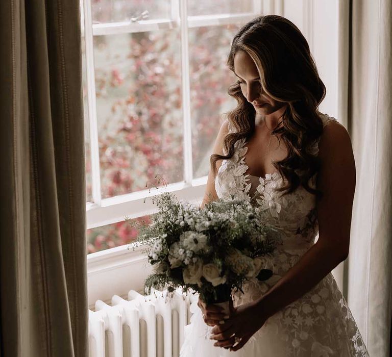 Bride stands at window as she finishes getting ready for her wedding with floral appliqué wedding dress and white bouquet 