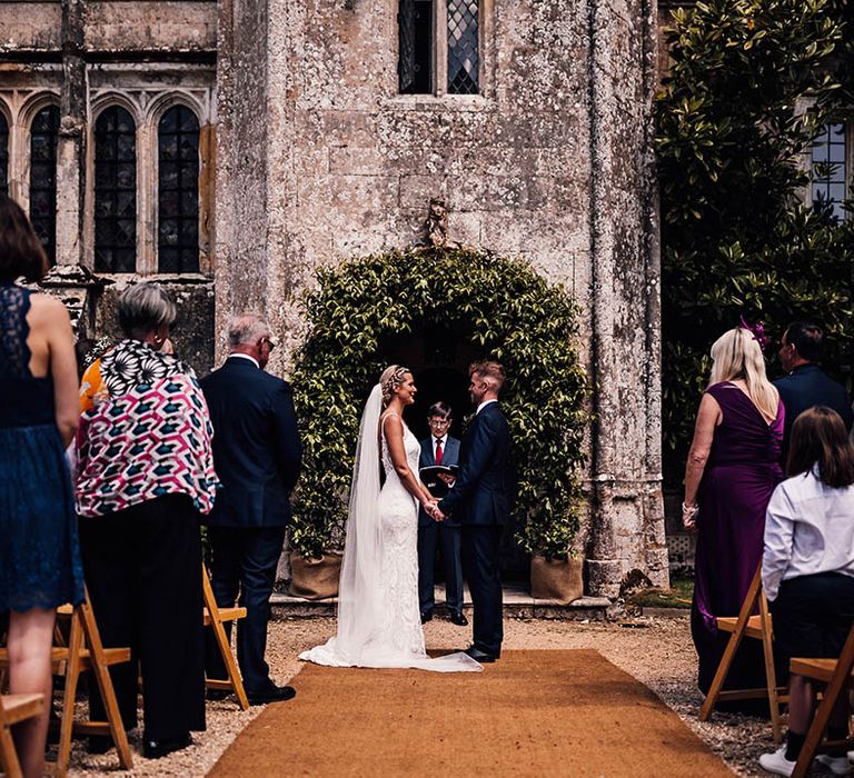 Bride and groom stand holding hands in front of foliage wedding arch for outdoor ceremony at Athelhampton House