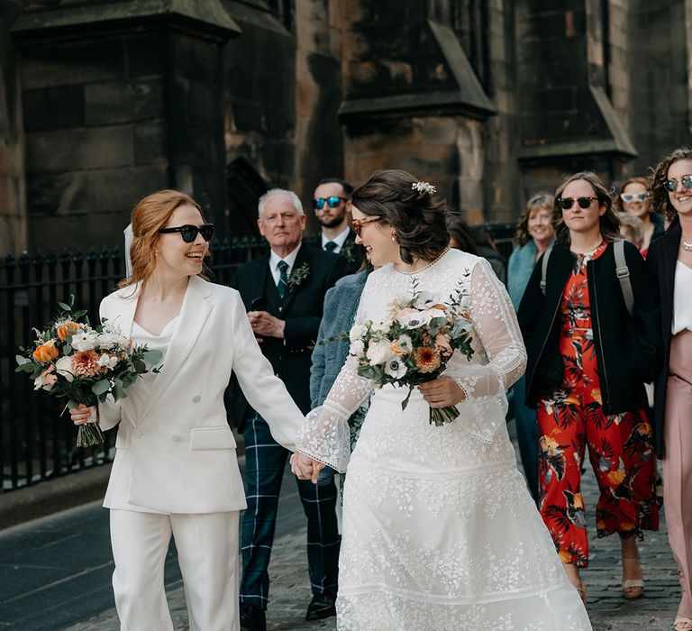 Two brides walk together in Edinburgh. One wears white bridal suit and the other wears boho gown from Coast, both carrying floral bouquets with pops of orange 