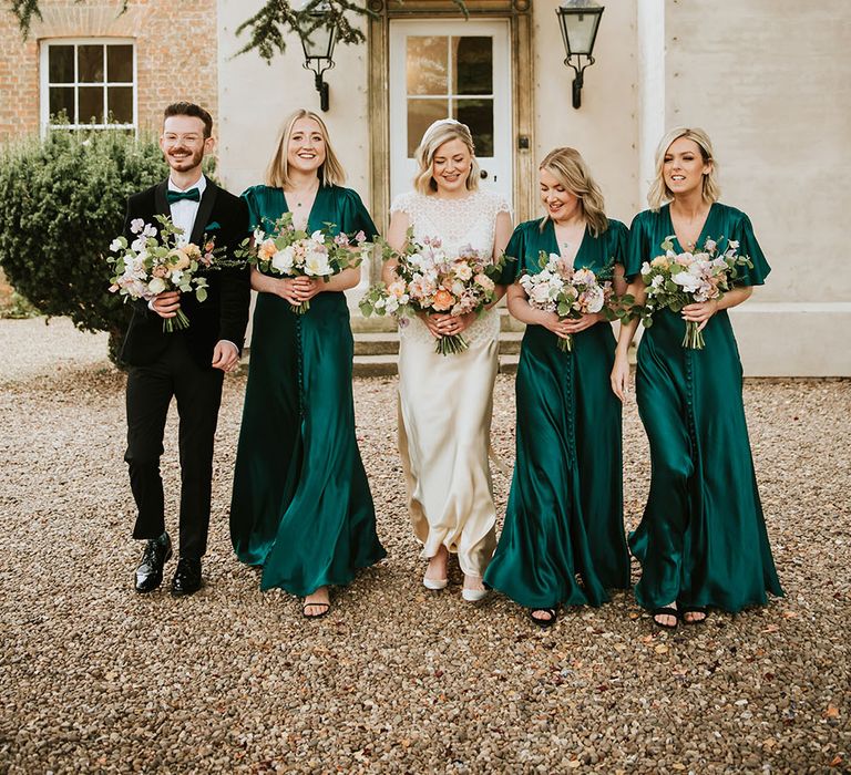 Bride stands with her Man of Honour and bridesmaids who wear green satin bridesmaids dresses with short sleeves and buttoned front