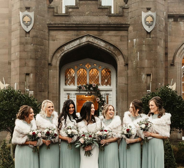 Bride stands with her bridesmaids who wear sage green high neck gowns complete with faux fur stoles and hold red and white floral bouquets