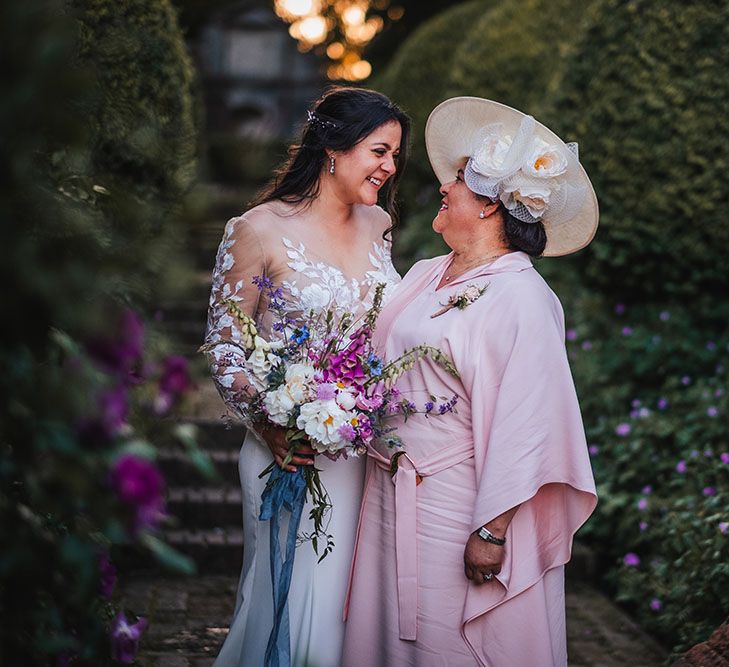 Bride wearing sheer lace embellished wedding dress looks lovingly at her mother who wears pastel pink dress and white hat with roses