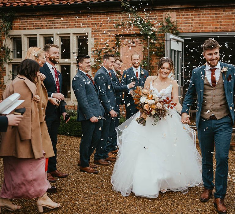 Groom in blue suit with brown waistcoat and burnt orange tie and handkerchief walks with bride in tulle skirt wedding dress as confetti is thrown