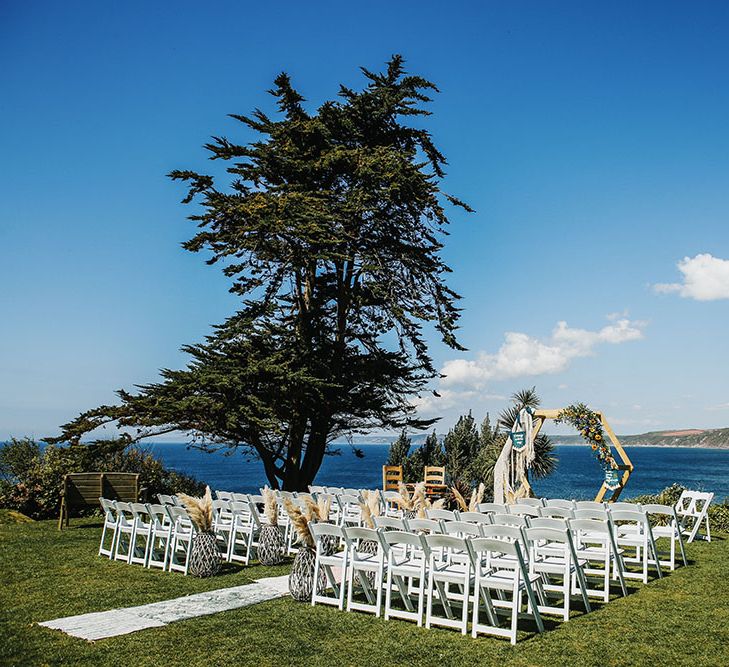 White chairs line aisle finished with Moroccan styled rugs and DIY wooden arch in front of the seafront in Cornwall  