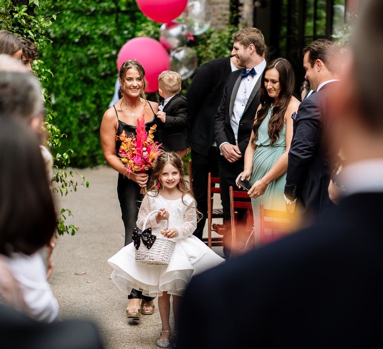 Flower girl in long sleeved ballerina styled dress walks with white wicker basket and bridesmaid in black silk bridesmaid dress