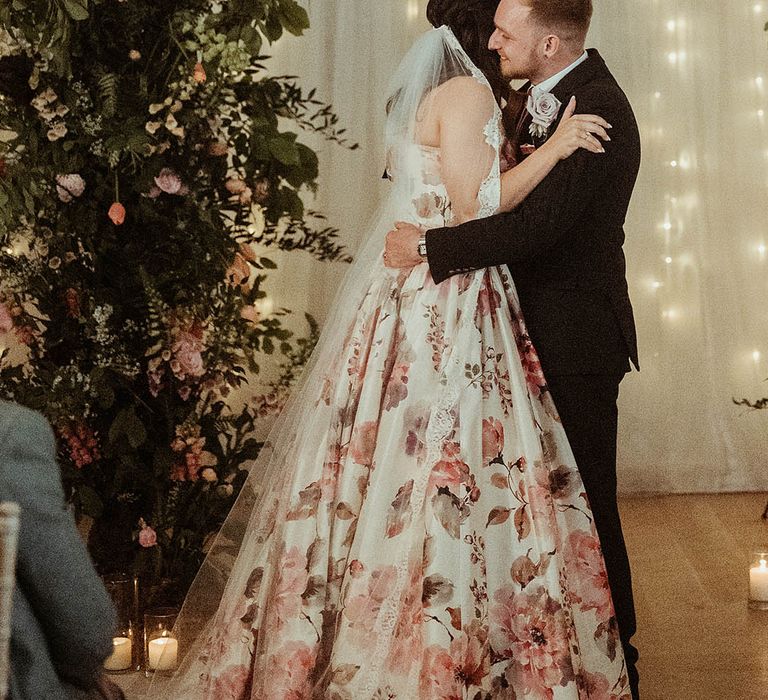 Bride in floral hand painted silk wedding dress hugs the groom at the altar 