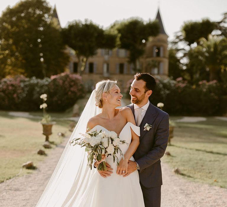 Bride wears Suzanne Neville fitted wedding dress with plunge neckline and off-the-shoulder detailing whilst carrying white rose floral bouquet 