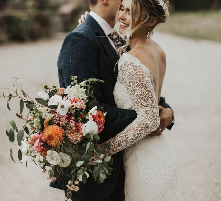 Bride in a backless lace long sleeve wedding dress with groom in a blue suit hugging the bride holding a peach, coral and white wedding bouquet 