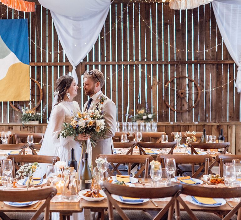 Bride and groom stand in their wedding reception at Deepdale Farm with colourful tapestries, bunting and table decor 