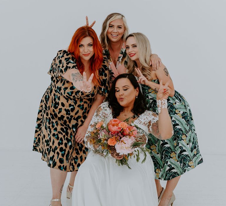 Bride holds brightly coloured floral bouquet and stands with wedding guests in brightly coloured outfits in front of white wall