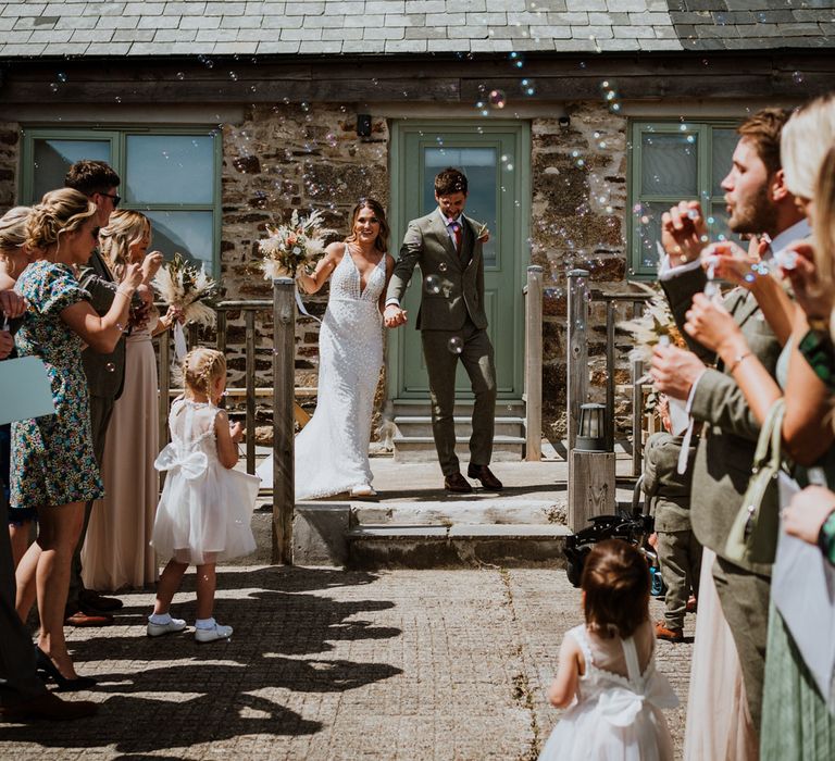 Bride in sleeveless Made With Love Bridal dress with white embellishments and groom in grey suit with orange pops of colour at The Green Cornwall doing a confetti bubble walk