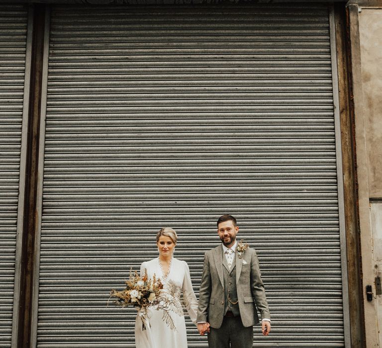 Groom wears three piece grey suit whilst stood beside his bride in Reformation wedding dress whilst holding dried floral bouquet 