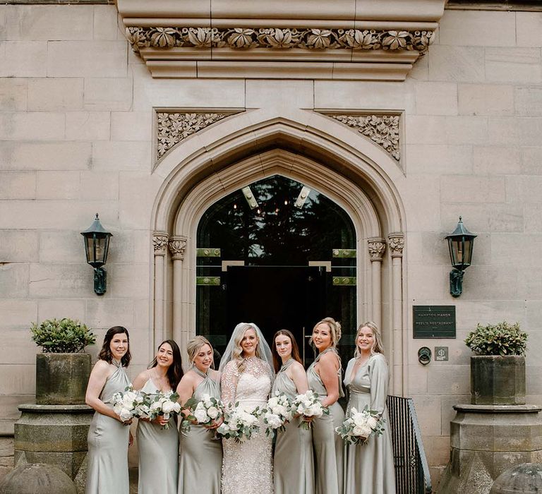 Bride in lace wedding dress stands with her bridesmaids in sage green satin bridesmaid dresses with different styling