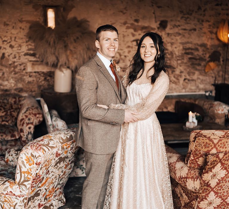 Groom wears brown suit complete with burnt orange tie as he stands with his bride in sheer gold wedding dress