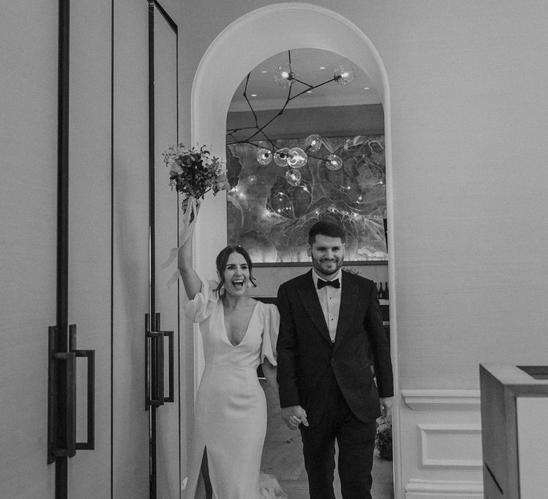 Bride in modern wedding dress with front slit and puff sleeves holding bouquet with groom in dark suit and large boutonniere 