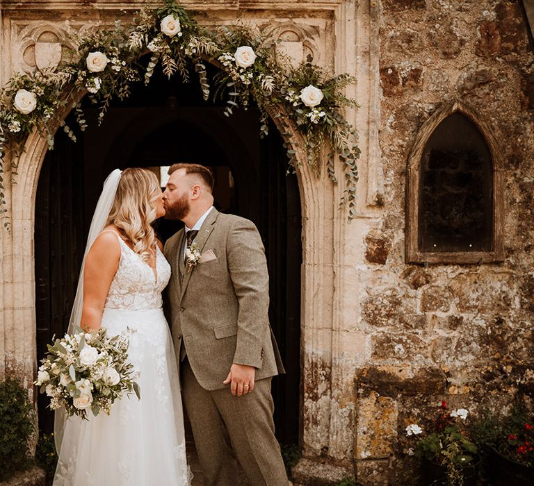 Bride in a lace wedding dress and groom in a grey suit share a kiss at the entrance to the church