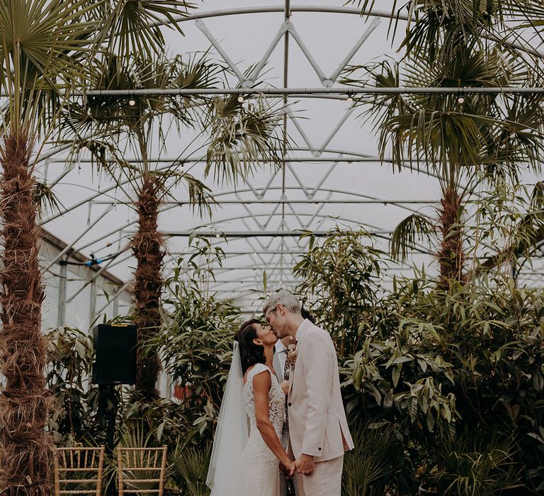 Bride in lace and tulle dress kisses groom in light pink suit in front of greenery