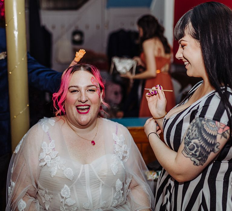 Bride in corset top wedding dress with lace overlay getting her face painted at wedding party