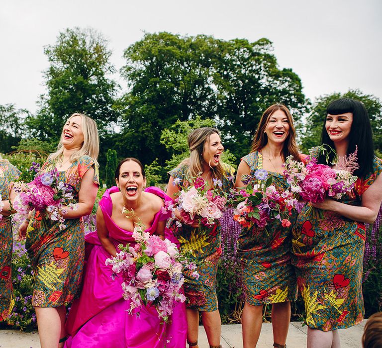 Bridesmaids in matching midi green colourful patterned Vivienne Westwood bridesmaid dresses with ruching, short sleeves standing and laughing with bride in off the shoulder front ruching pink Vivienne Westwood wedding dress holding pink floral bouquet with pink peonies, fuchsia roses, lilac sweet peas and green foliage