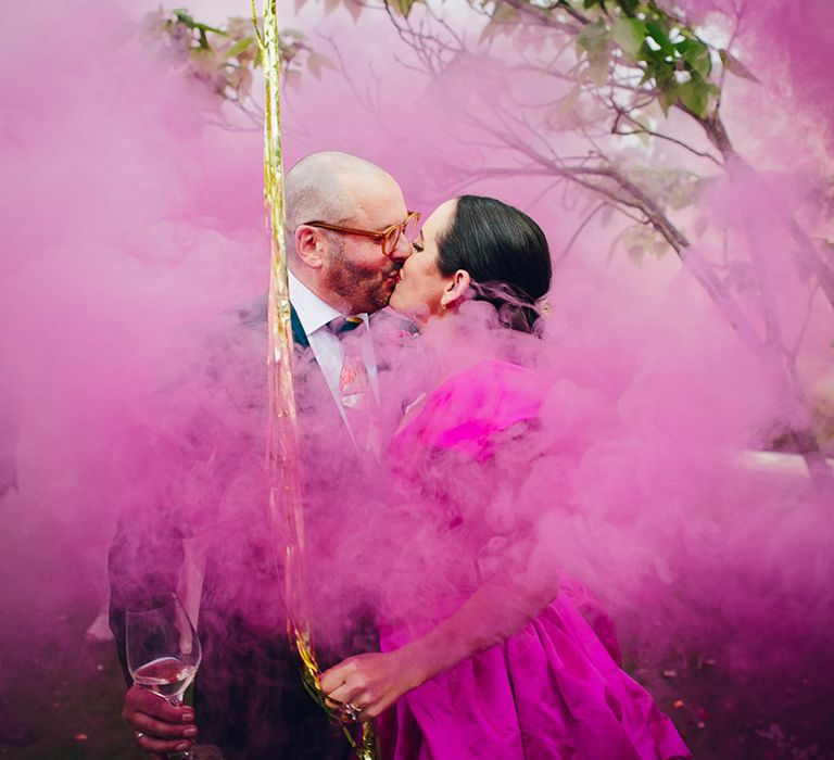 Bride and groom holding champagne doing bright pink smoke bomb photoshoot, bride holding onto gold streamers