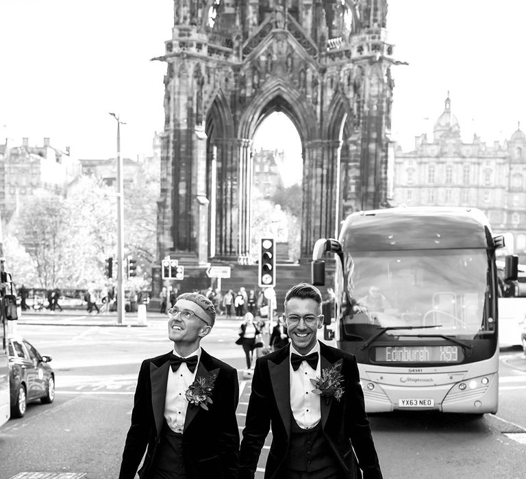 Grooms in black velvet tuxedos, white shirts, black bowties, and rose and foliage boutonnieres walking hand in hand through Edinburgh city centre