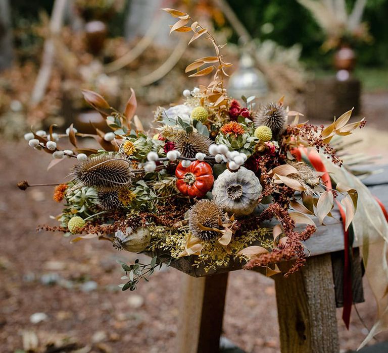 Dried autumnal wedding bouquet with white dried cotton stems, terra cotta celosia seeds, green foliage and pumpkins, gourds and fruits from the trees on a wooden stool at a harvest wedding