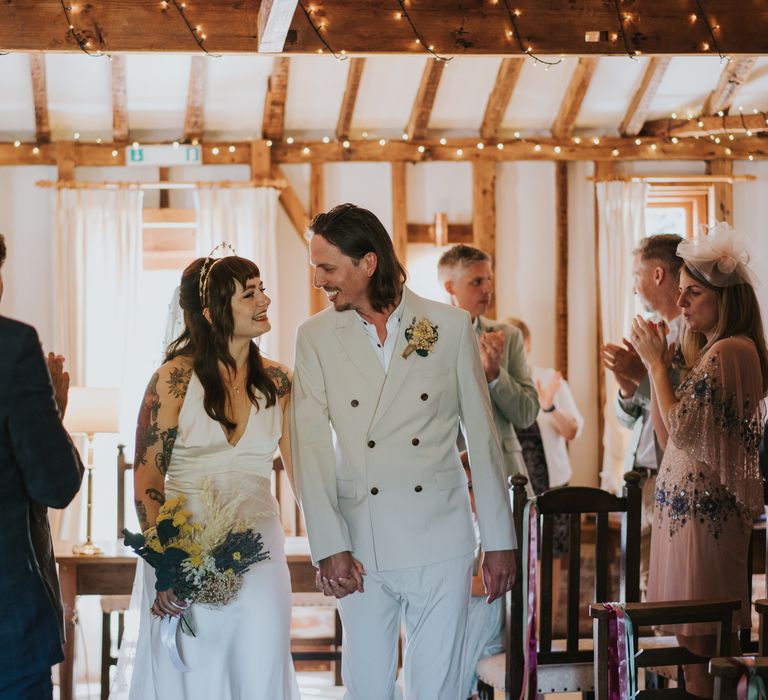 Bride holding dried flower wedding bouquet with white bunny tails, green foliage, yellow dried gypsophila, lavender and baby’s-breath, smiling at groom wearing neutral coloured double-breasted ASOS suit and black high-top converse with dried flower and foliage boutonniere after wedding ceremony