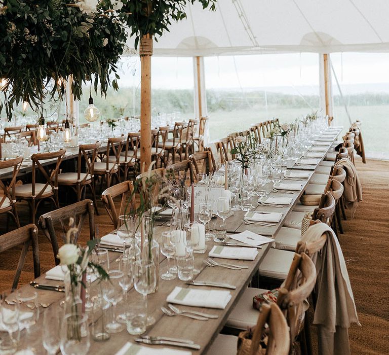 Marquee wedding reception complete with wooden banquet tables, green foliage installations hanging above and wicker lights