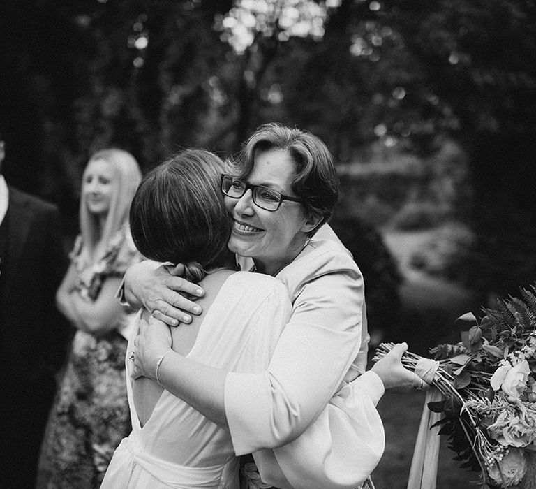 Wedding guest hugs bride outdoors whilst holding floral bouquet 