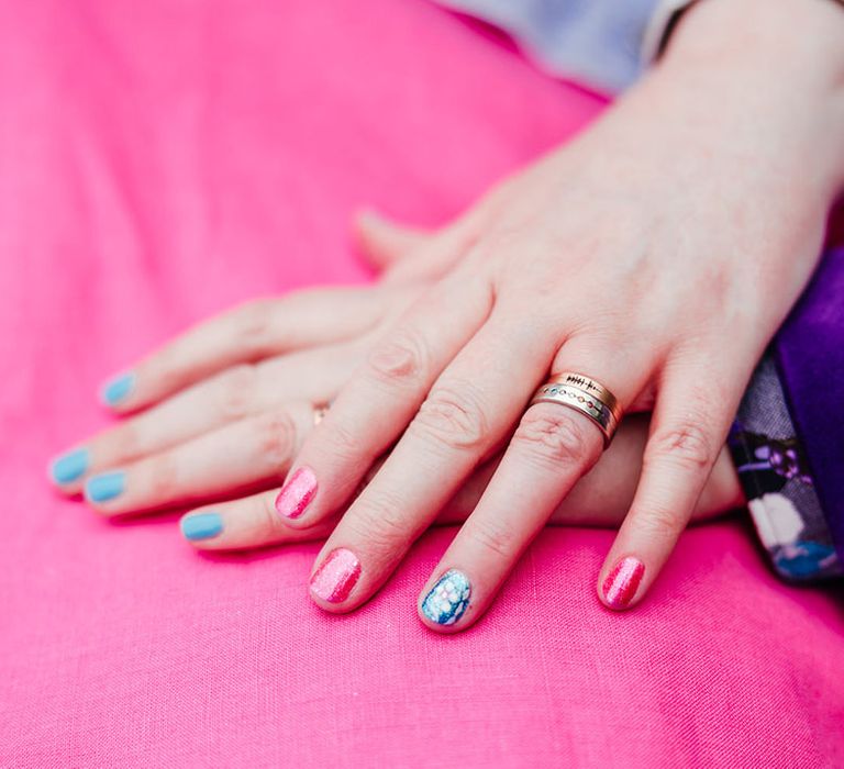 Brides with complimenting sparkly pink and blue nail varnish showing off their wedding rings in each other's hands on top of pink wedding dress with the sleeve of a purple velvet blazer 